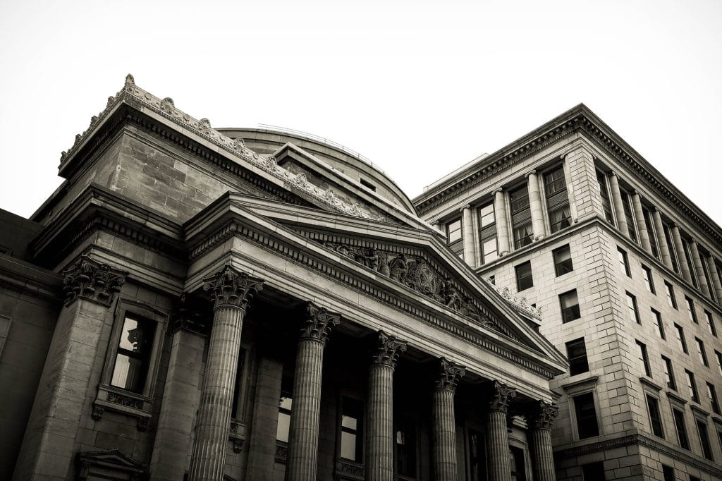 Imposing grey stone buildings loom at an odd angle from above. The architecture of one of the buildings (columns and a triangle frontage) infer it is a banking institution. 