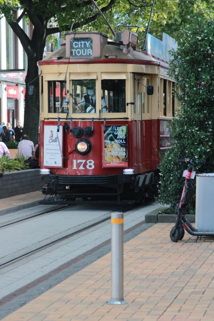 The front of an old maroon and beige city tram appears in a city scape with the words “City Tour” displayed on it. A famous presence in Christchurch city centre, the tram passes near Christchurch Casino. 