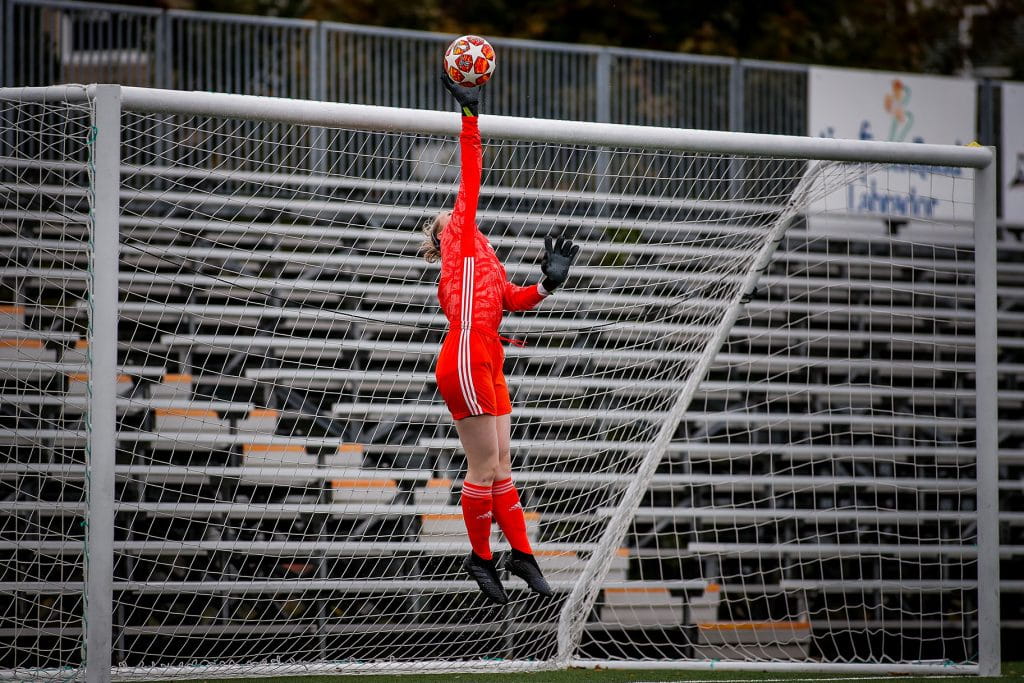 А woman in a red soccer team kicks a soccer ball over the goal net. 