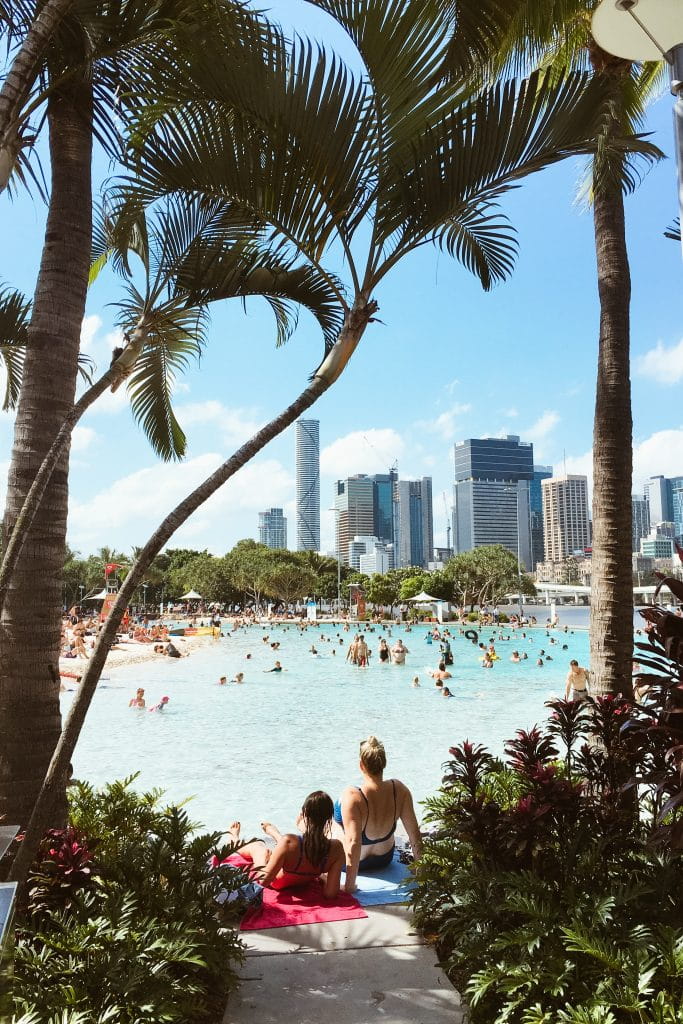 A beautiful lagoon with yellow sandy beaches is framed by palm trees. Modern inner-city skyscrapers create a backdrop on the horizon. 