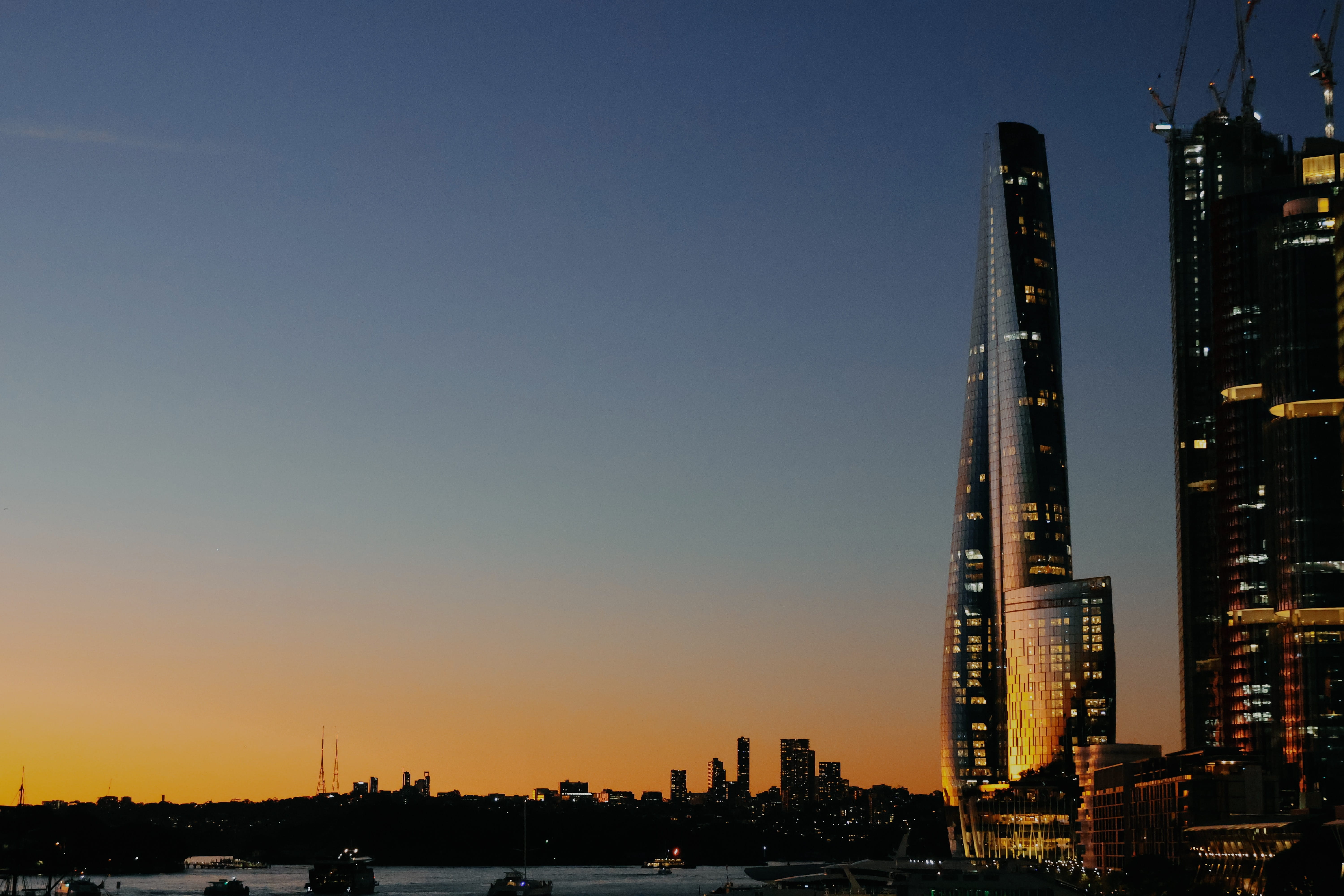 The sky graduates from blues to oranges as the sun sets over Sydney Harbour. The orange horizon is reflected by the window-clad building, Crown Sydney, which contains a VIP-only casino.