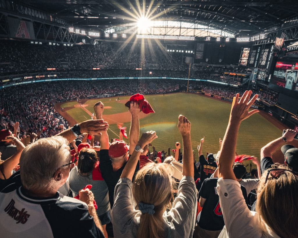 A giant stadium is completely filled with cheering people. A baseball diamond can be seen, as if from the top section of the tall bleachers. In the foreground fans stand and wave their arms, wearing or waving red-coloured items. The mood is ecstatic.