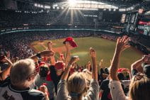 A giant stadium is completely filled with cheering people. A baseball diamond can be seen, as if from the top section of the tall bleachers. In the foreground fans stand and wave their arms, wearing or waving red-coloured items. The mood is ecstatic.