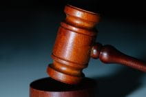 A brown-stained wooden gavel rests on a greyish-blue surface. The shadow in the background gives an ominous and serious feel.