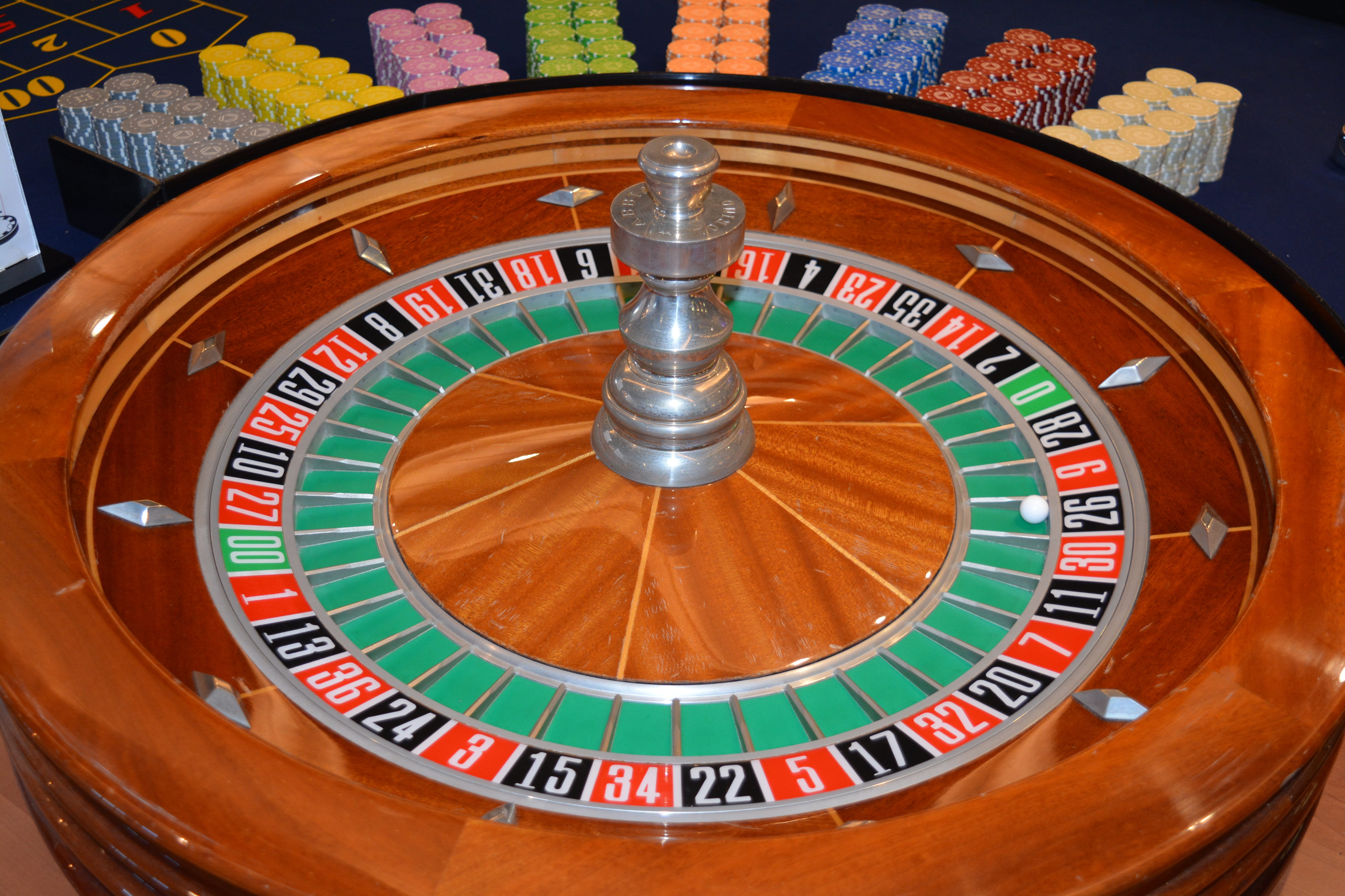 A beautiful varnished wooden roulette wheel sits in the foreground. Eight different colours of tokens are stacked up in rows behind it on a deep blue velvet-covered table. The white ball sits at number 26.