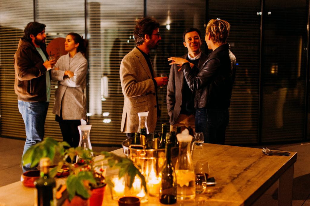 In an ambiently lit modern room, two groups of people network at an evening event. They are engaged and energised. The interior design of the room is modern and professional but warm.