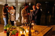 In an ambiently lit modern room, two groups of people network at an evening event. They are engaged and energised. The interior design of the room is modern and professional but warm.