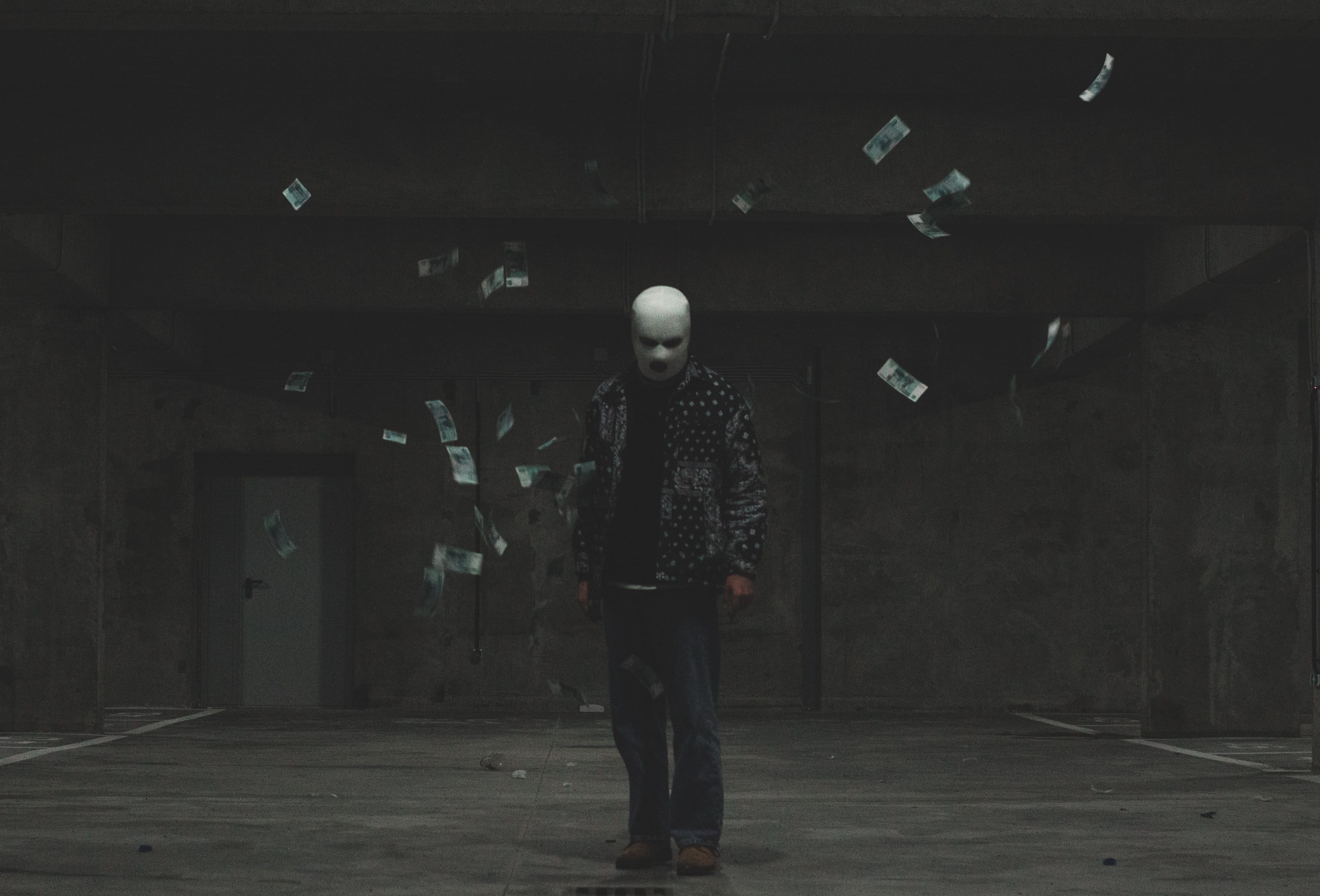 A man in a white balaclava stands in a dark concrete-walled room. Cash is flying through the air around him. He is wearing tough street clothes - a black jacket with squarish dots that emulate studs.