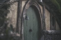 A green oval door and grass hill roof of a Hobbit’s home. It has a weathered wooden architrave and beside it is a blackened bell. A small lamp hangs above the door. Together with a white picket fence, it feels comfortable and homey.