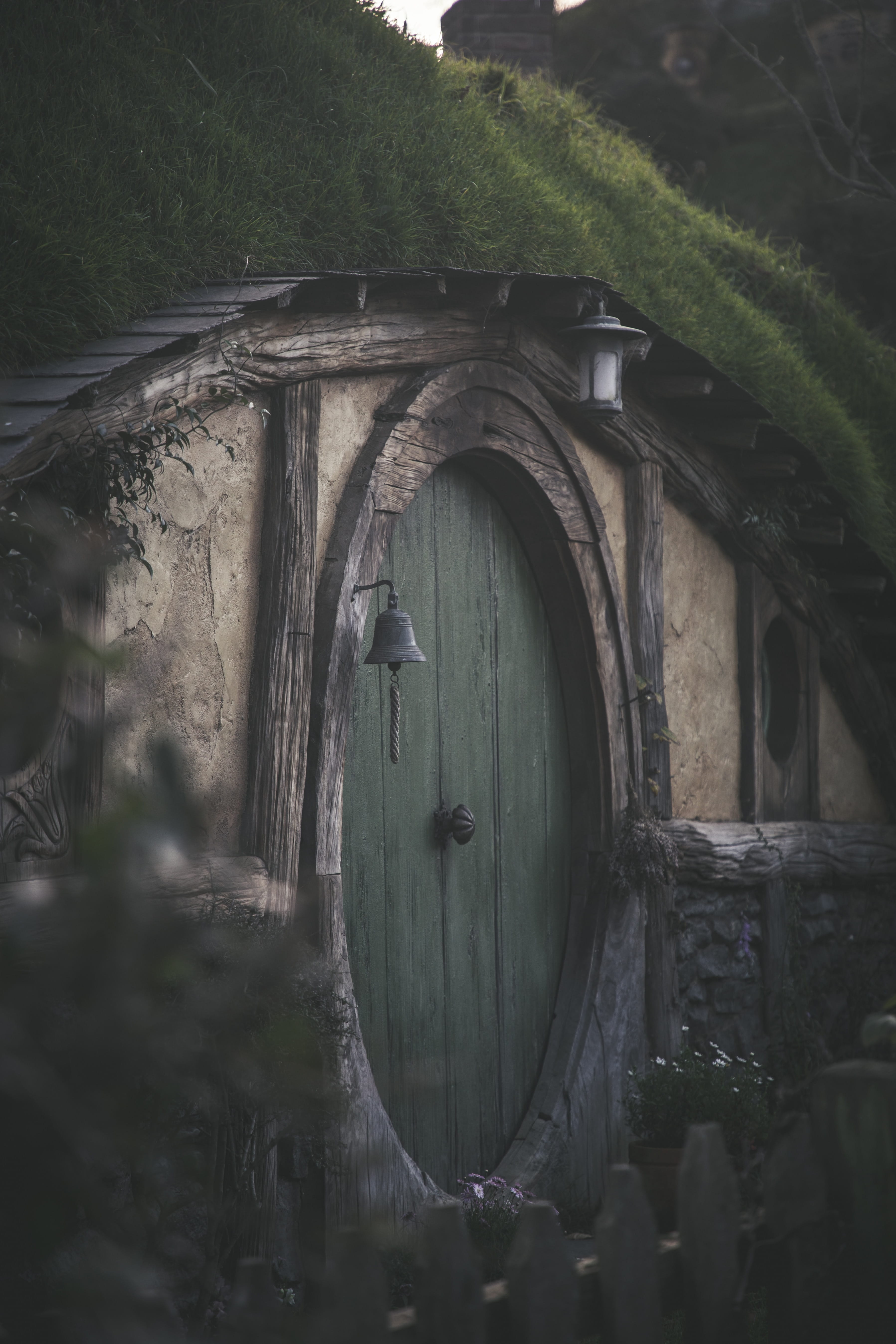  A green oval door and grass hill roof of a Hobbit’s home. It has a weathered wooden architrave and beside it is a blackened bell. A small lamp hangs above the door. Together with a white picket fence, it feels comfortable and homey.