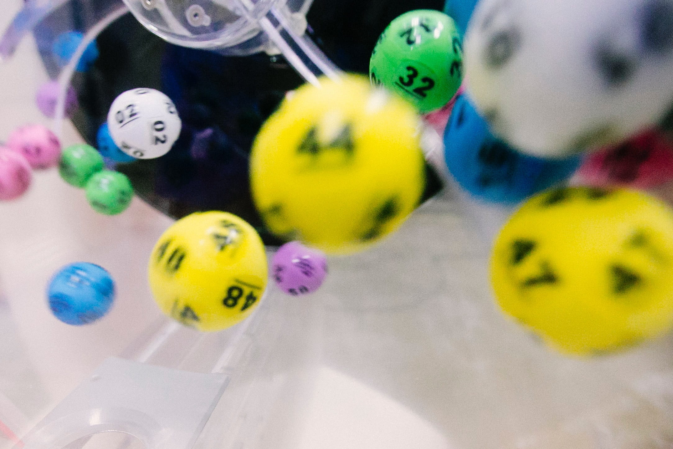 Two dozen balls are in mid-air, bouncing around inside a clear plastic lottery machine. Bright yellow balls are prominent, with their numbers printed on them. A green ball is clearly marked, “32”.