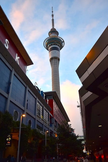 A triangle of blue sky can be seen between two buildings. Auckland’s skinny SkyTower reaches up to the clouds from between these two long buildings. The one on the left displays large separate neon letters on its frontage. They spell out SkyCity.