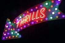 A giant arrow sign contains the word “thrills” in all caps. Coloured light bulbs in red, purple, yellow and white decorate the sign. The word is in white neon with a red colour behind it.