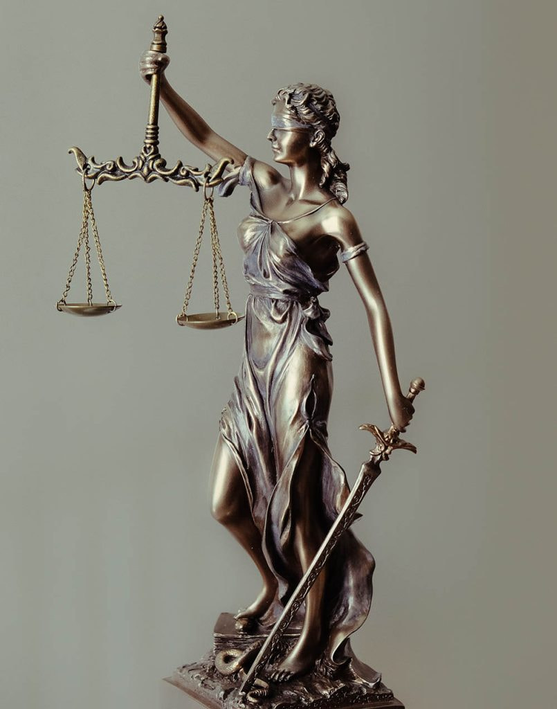 A metal statuette of Lady Justice stands with a grey background. She is holding up scales in one hand, and in the other, a sword pointed at the ground. She is blindfolded. 