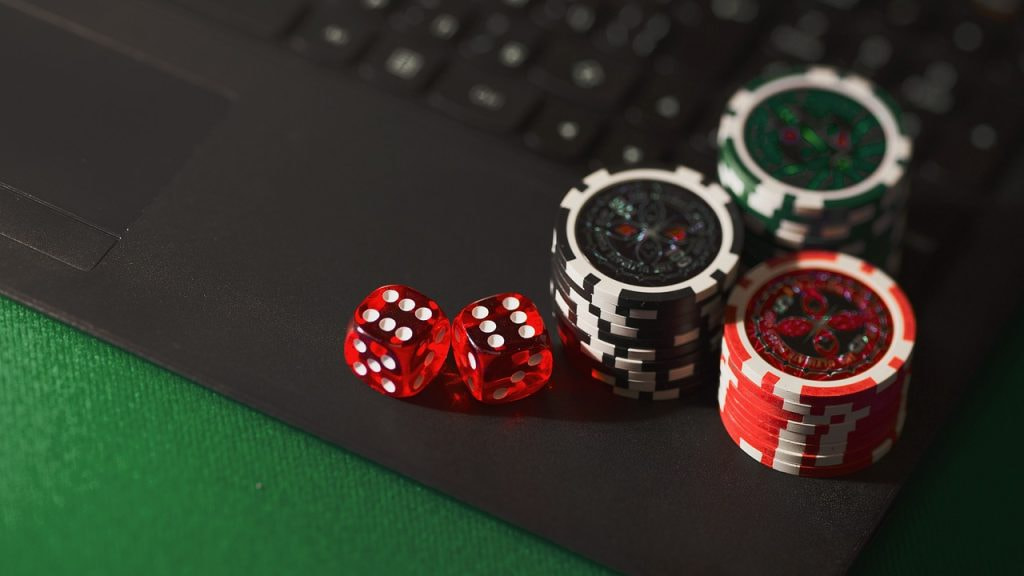 A black laptop sits on top of green material. The green is a similar shade to that which is found on gambling tables at casinos. Three stacks of gambling chips and a pair of dice sit on the edge of the laptop.