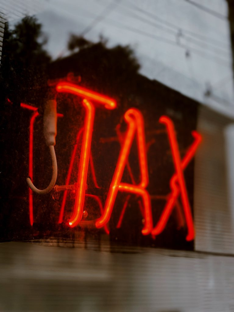 A neon sign blazes red on a reflective black background, the word “TAX” in all-caps. 