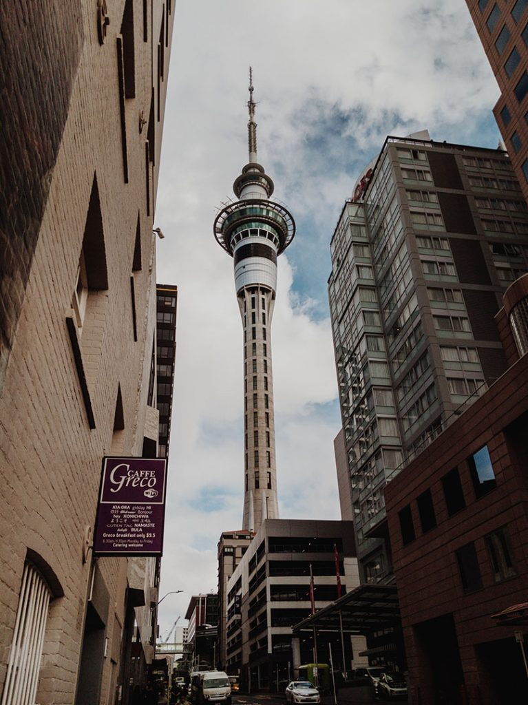 Sky Tower reaches up into a cloudy sky, looming above the street buidings on either side of the road. Some cars are parked on the street. 