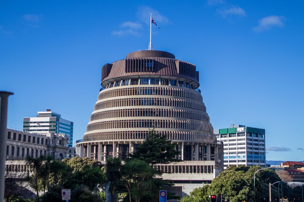 The New Zealand Parliament building, shaped like it’s common name, The Beehive, stands tall in front of a bright blue sky. Some trees can be seen, small at its base. A few shorter buildings surround it.