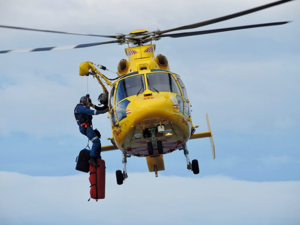 A bright yellow helicopter, with the red and white checkers of emergency services beneath its blades, hovers. A man, strapped to a line, is getting ready to descend from outside the helicopter with two bags of supplies.