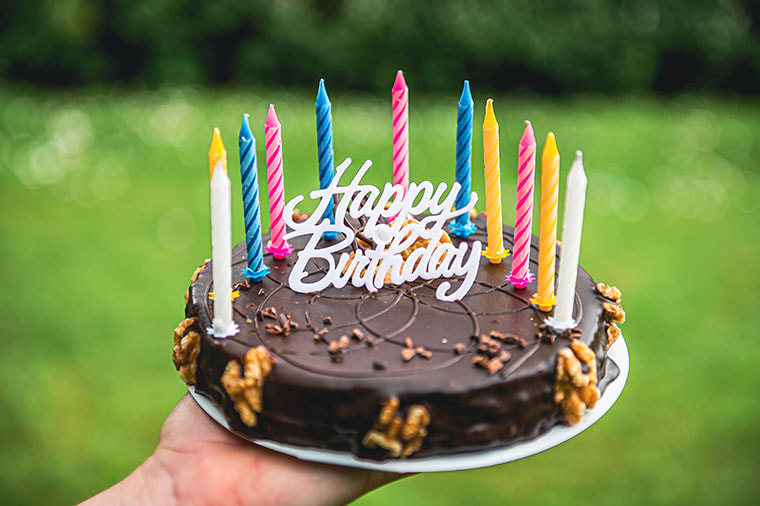 A chocolate birthday cake is held out in front of a background of greenery. Yellow, white, pink and blue birthday candles surround a white plastic cake topper that says, “Happy Birthday” in elegant running writing. 