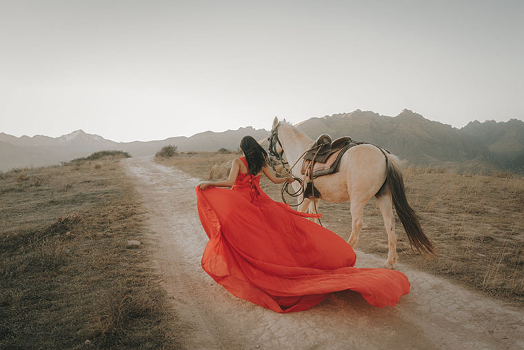 A saddled white horse is led by its reins along a mountain path by a woman in a red dress. The dress is very long and billows out around and behind her. 