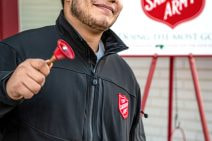 A young man rings a small red bell. He wears a black jacket with a Salvation Army badge sewn on its front. A Salvation Army sign with a collection bucket hanging from it is behind him.