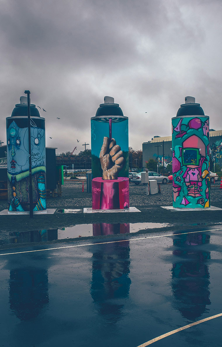 Three statues stand beside a bitumen road. The sky is cloudy and grey behind them. The statues are each in the shape of spray cans and showcase graffiti and street art on their sides. 