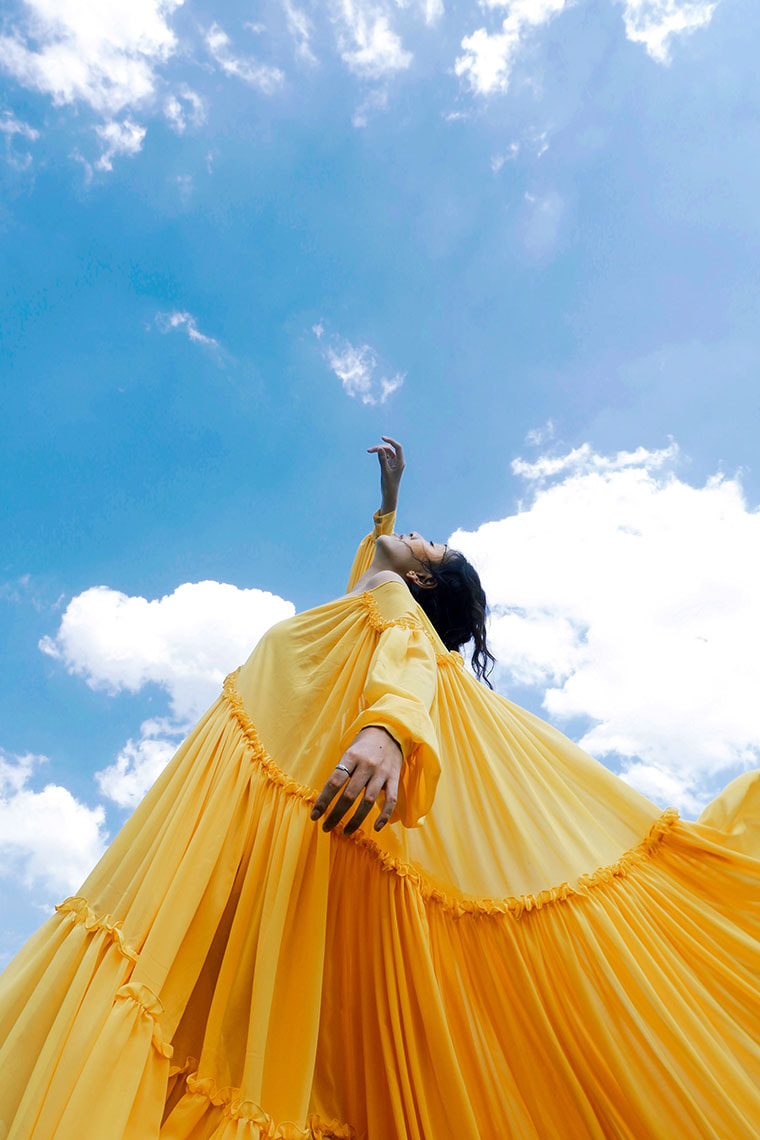 A woman in a yellow dress reaches up to the bright blue sky with one hand in a ballet pose. She is wearing a fashion designer dress; it has a huge amount of fabric, layers of rouching and small frills in between each layer.