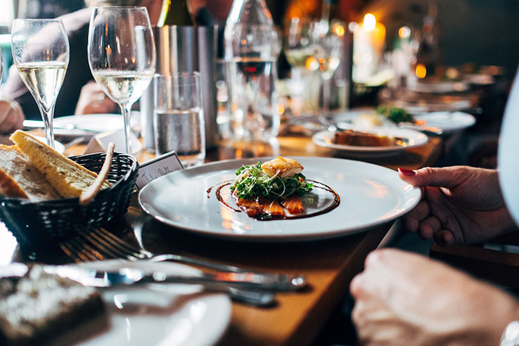 A long wooden table is full to the brim with plates, glasses and cutlery. A pale blue ceramic dinner plate hosts an expensive-looking modern course of fish and salad. A manicured hand is touching the plate and a basket of bread is behind it.